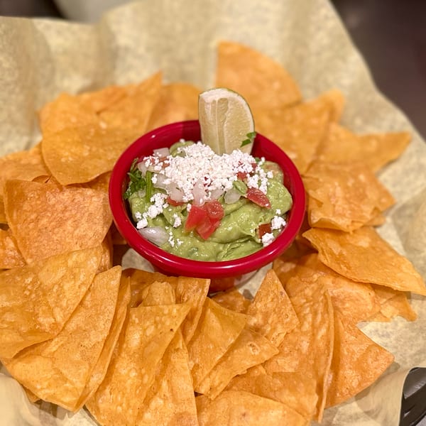 A red bowl of guacamole topped with cheese, tomatoes, and a lime wedge, surrounded by tortilla chips.