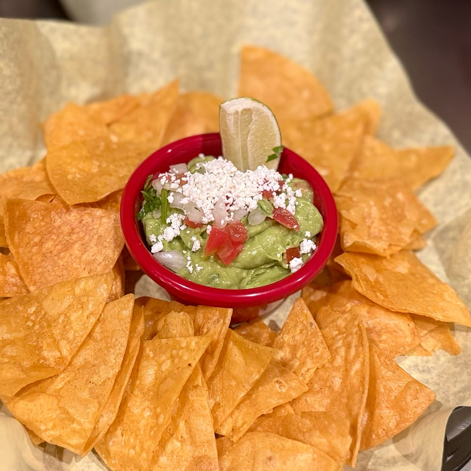 A red bowl of guacamole topped with cheese, tomatoes, and a lime wedge, surrounded by tortilla chips.