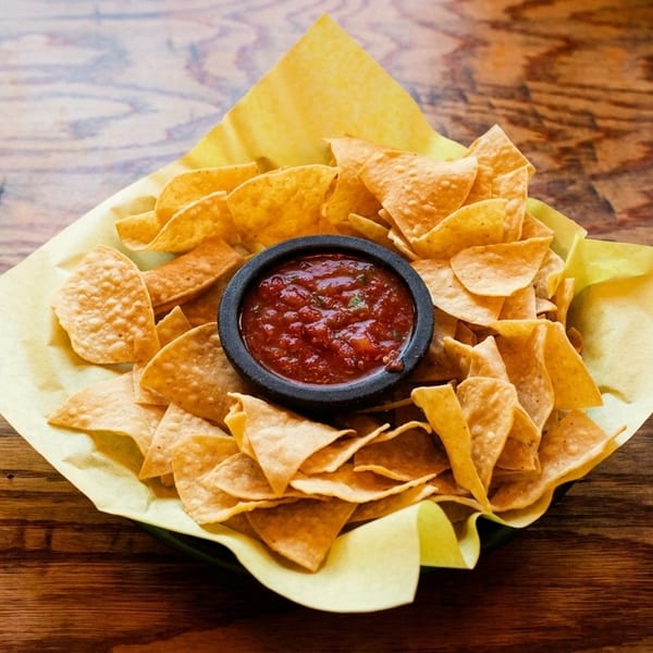 A bowl of tortilla chips with a small black bowl of salsa in the center, on a wooden table.