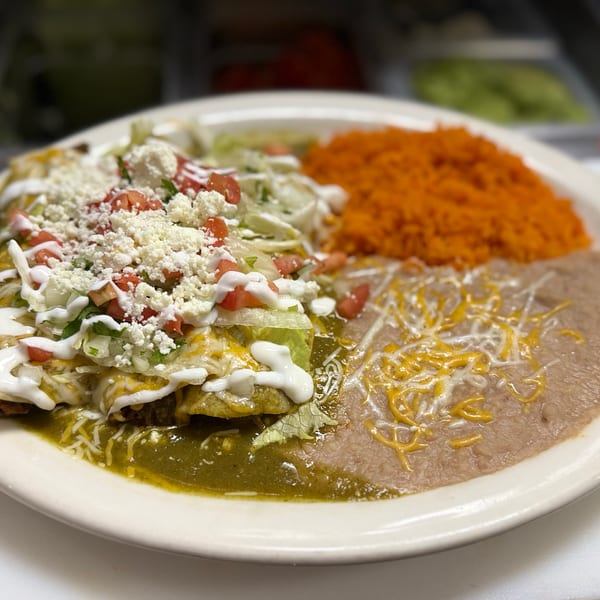 A plate of enchiladas verdes with rice and refried beans, topped with cheese, lettuce, and tomatoes.